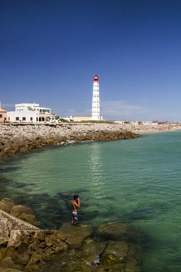 Lighthouse by rocky shore and swimmer in water.